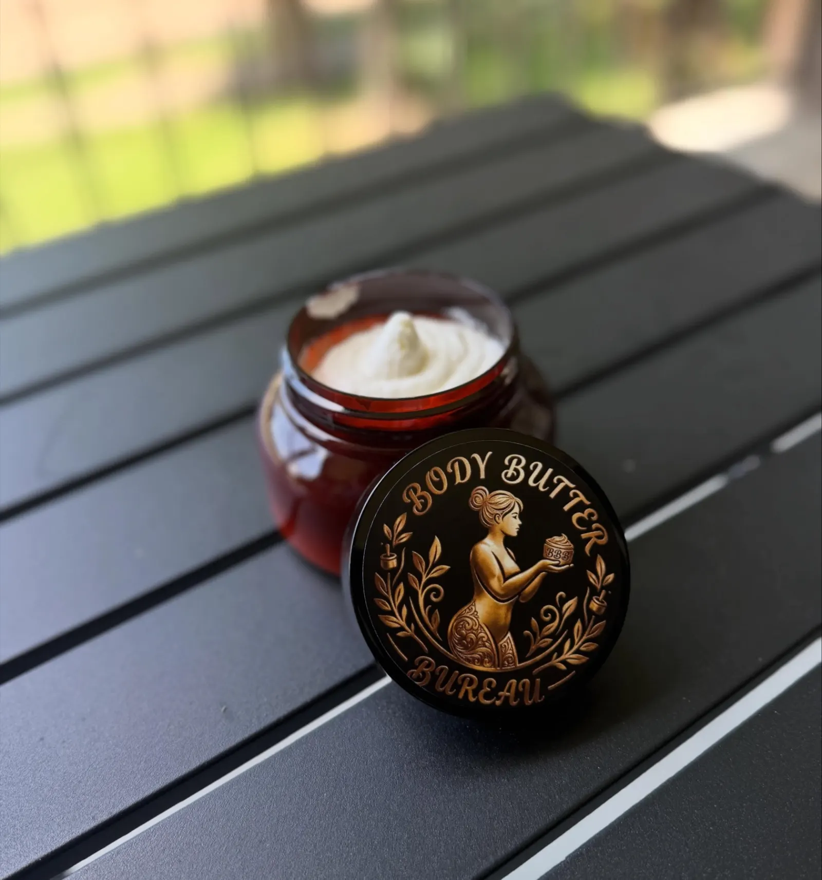 An open amber jar of whipped shea butter with a soft peak, beside its black lid with the gold Body Butter Bureau seal