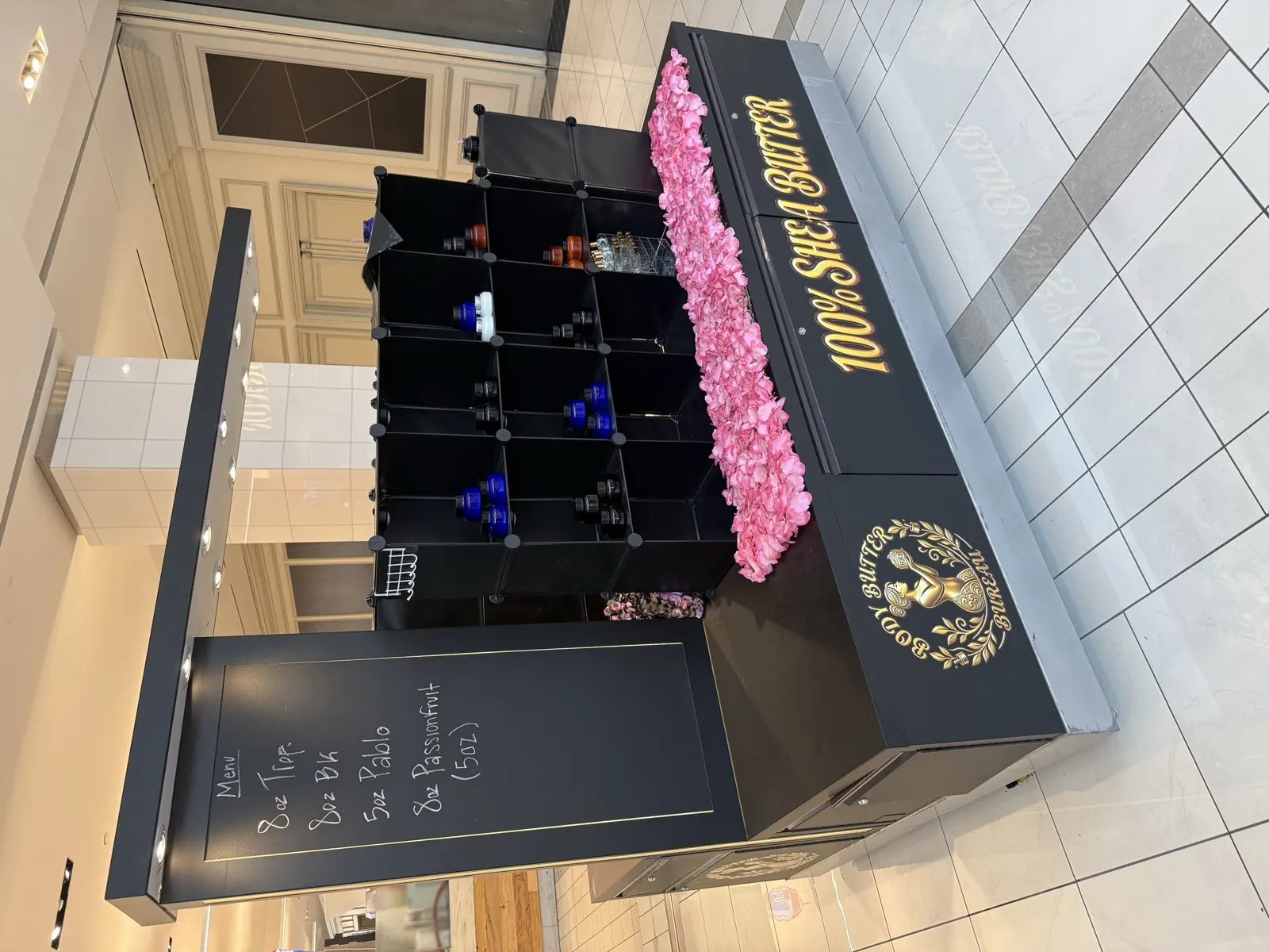 The black and gold Body Butter Bureau kiosk with pink petals and a chalkboard menu